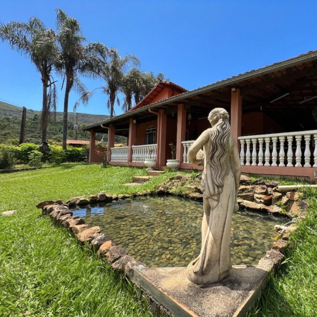 Área externa do Recanto do Bem em Moeda, MG, com lago ornamental de pedras, estátua decorativa e vista para a varanda da casa de campo rústica.