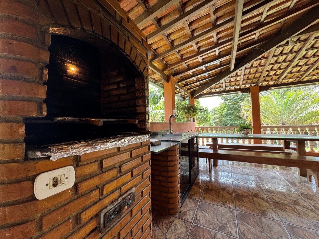 Área gourmet rústica com churrasqueira de tijolinho à vista, bancada em granito, mesa de madeira longa e vista para o jardim no Recanto do Bem em Moeda