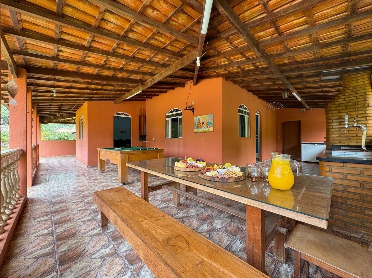 Varanda gourmet ampla com mesa rústica de madeira posta com tábuas de frios e suco natural, mesa de sinuca ao fundo, churrasqueira de tijolinho e chopeira no sítio Recanto do Bem em Moeda.