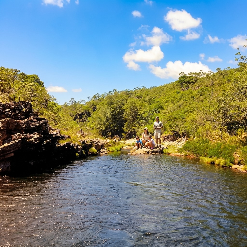 Fotografia de uma piscina natural de águas escuras no primeiro plano, com reflexos do céu azul e nuvens brancas. Ao fundo, uma família de quatro pessoas (mãe, pai e dois filhos) está parada sobre pedras em um poço raso. Uma cachoeira suave cai atrás deles entre grandes rochas. O cenário é cercado por uma densa floresta verde sob um céu azul ensolarado.