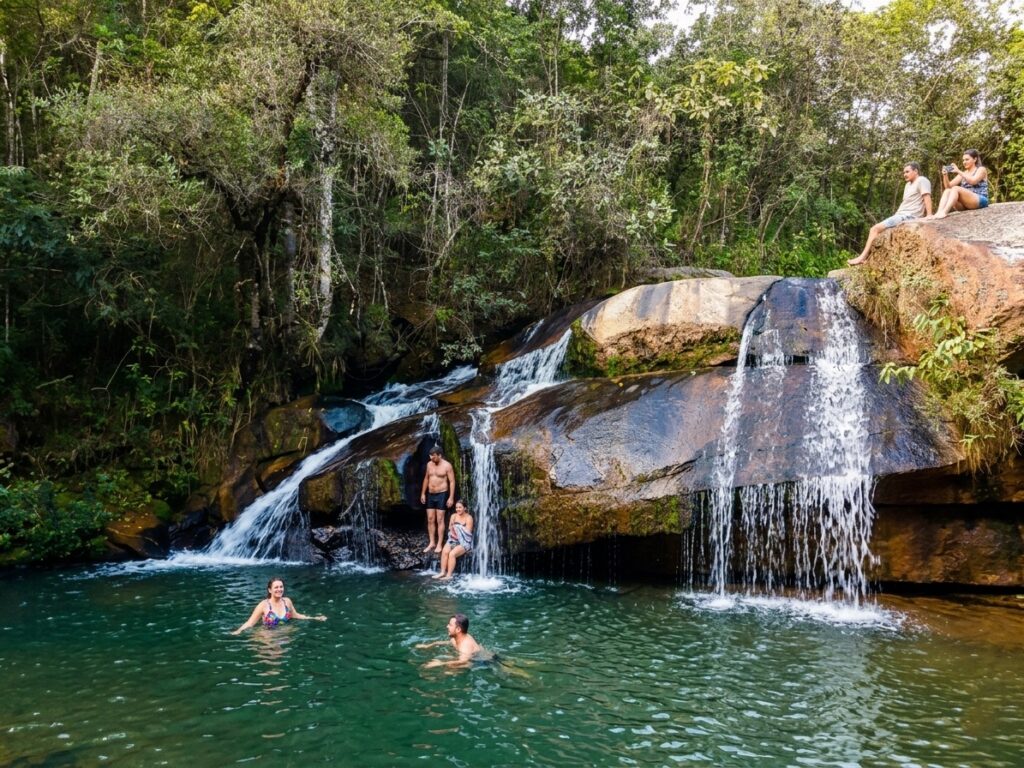 Grupo de amigos se refrescando em cachoeira de águas cristalinas na Serra da Moeda, MG. Ao fundo, mata preservada e quedas d'água sobre pedras naturais.
