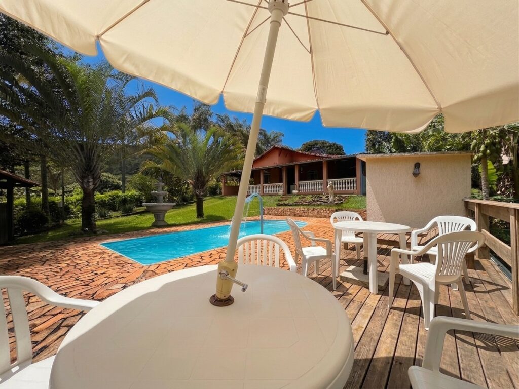 Área de lazer do Recanto do Bem com deck de madeira, mesas sob guarda-sol e piscina ao fundo cercada por jardim tropical.