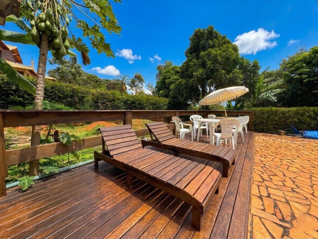 Deck de madeira revitalizado com espreguiçadeiras sob um céu azul ensolarado, área de mesas com guarda-sol e vista para a vegetação no sítio Recanto do Bem em Moeda.