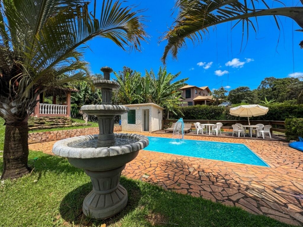 Área de lazer da casa de campo Recanto do Bem em Moeda MG, com piscina, cascata e jardim com chafariz sob céu azul.