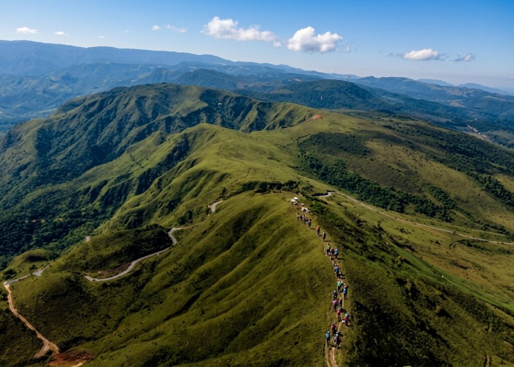 Vista panorâmica da Serra da Moeda em um dia ensolarado, mostrando a trilha para a rampa de voo livre com um grupo de trilheiros e parapentes no céu azul.