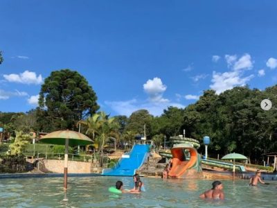 Crianças e adultos se divertindo na piscina artificial do balneário da Cachoeira do Moinho, em Moeda, MG, com toboáguas e cercada por árvores e céu azul.