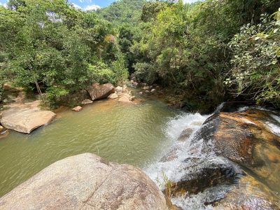 Vista de cima da borda da Cachoeira do Paiolinho em Moeda, Minas Gerais, mostrando o fluxo de água caindo em um poço esverdeado cercado por pedras grandes e vegetação nativa densa.