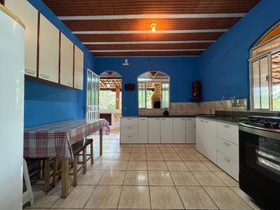 Cozinha interna ampla com armários brancos, fogão, geladeira e mesa de jantar, apresentando paredes azuis e vista para a área gourmet externa no Recanto do Bem.