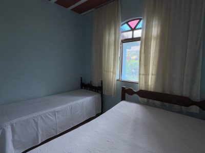 Quarto familiar iluminado com uma cama de casal e uma cama de solteiro extra, decorado com colchas brancas trabalhadas e janela com vitral colorido no Recanto do Bem.